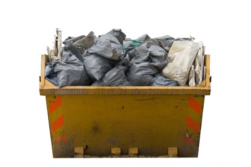 A large yellow skip bin filled with a variety of rubbish bags and debris, situated outdoors likely on a driveway or street. The bags are predominantly dark grey, with some white and green plastic packaging visible. The rubbish appears bulky and loosely packed, with some bags leaning or stacked atop each other. The skip has red and orange reflective safety markings on its sides and shows signs of dirt and wear on its surface. The background is plain and white, emphasizing the focus on the skip and its contents. This scene is typical of a rubbish removal service in Sydenham, demonstrating the type of waste collection handled by Flat Clearance Sydenham, with the skip ready for loading or collection in an urban or suburban setting.